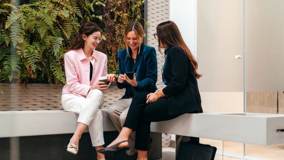 co-workers chatting during a work break at their office, one of them shows her friends something funny on her phone
