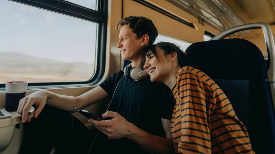 Happy young couple with phone and headphones looking out the window of a train