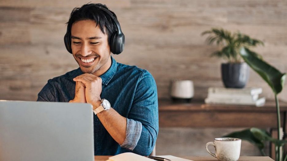 Smiling man with headphones in a denim shirt is looking at his laptop 