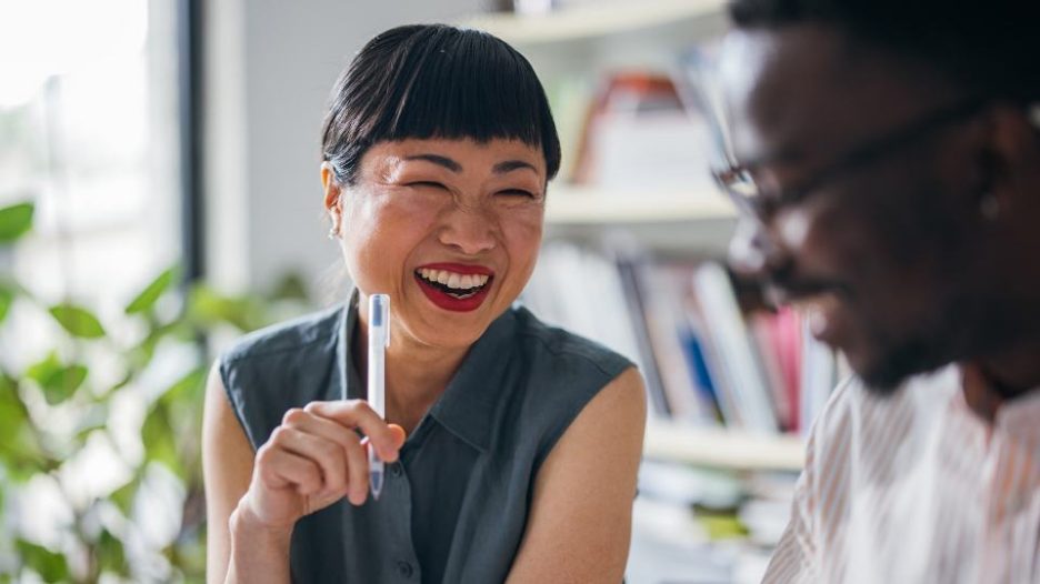 A beautiful Japanese entrepreneur laughing while holding a pen and talking to her coworker. (selective focus)
