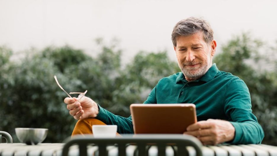 senior man sitting outdoors on terrace, drinking coffee and using digital tablet