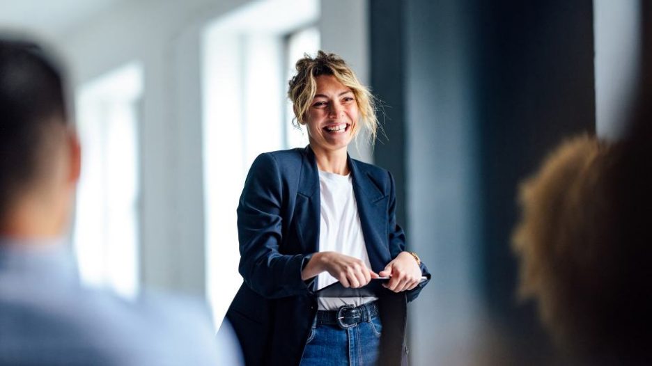 Cheerful woman in a dark blue jacket standing in front of a crowd looking down at a tablet.