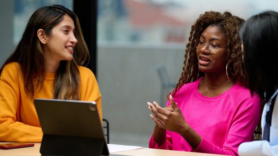 woman in casual clothes with braids and glasses talking about information on tablet to female coworkers 