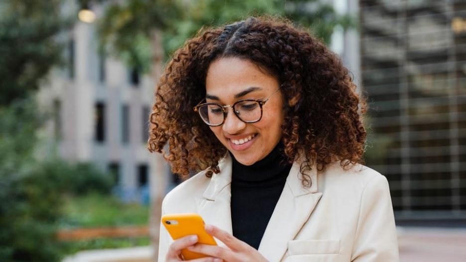 portrait of smiling businesswoman using smartphone in front of office building during a break from work
