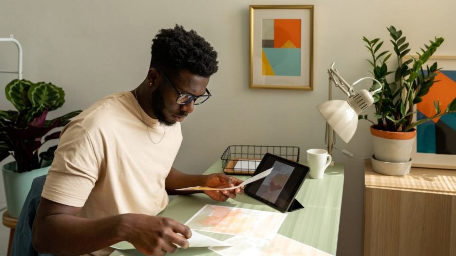 male in t-shirt working from office looking at images on sheets of paper