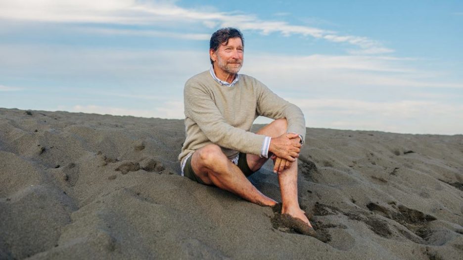 mature man smiling as he sits on a sandy beach
