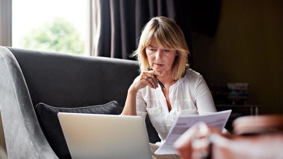 Pensive woman on a grey sofa looking at her laptop and paperwork