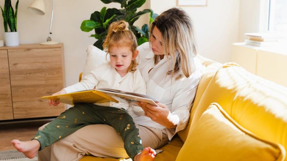 mum and young daughter reading a book and doing school homework sitting on yellow couch at home