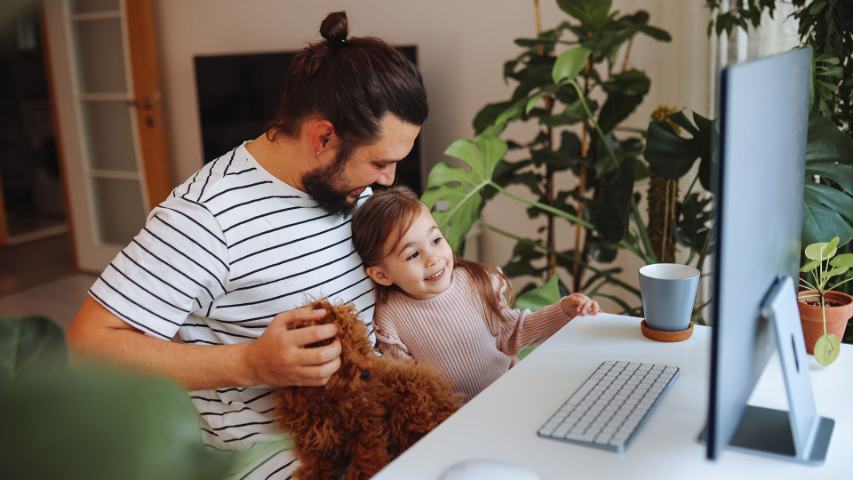 Father with young daughter and a puppy sitting on his lap in front of a computer in a modern sitting room