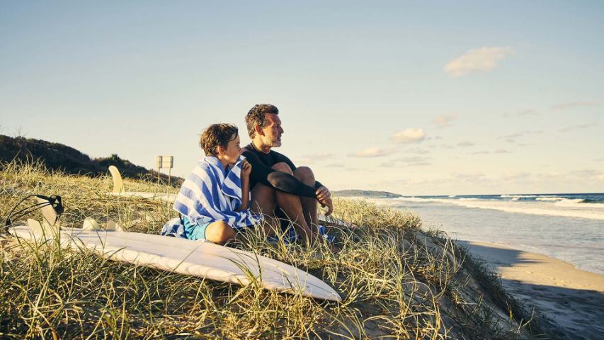 Father and son sitting in the dunes with a surfboard looking out to sea