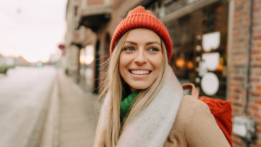 smiling woman in a scarf with an orange beanie is on the sidewalk outside a cafe