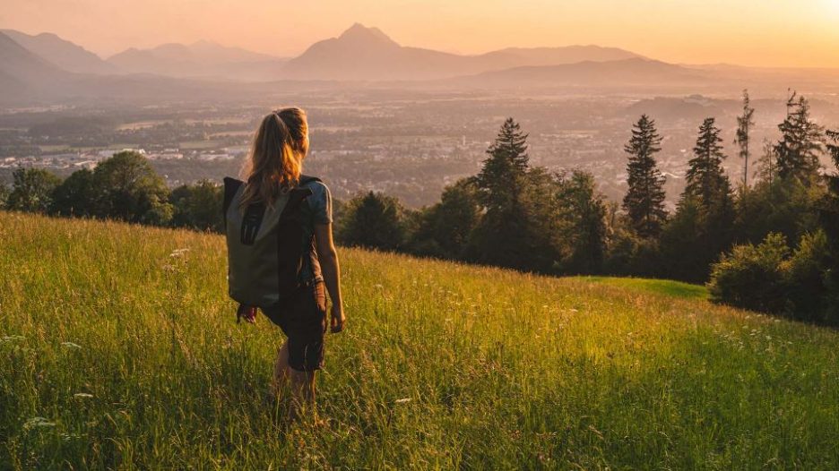 A woman with a backpack is hiking through a field at sunset with a city and mountains in the distance