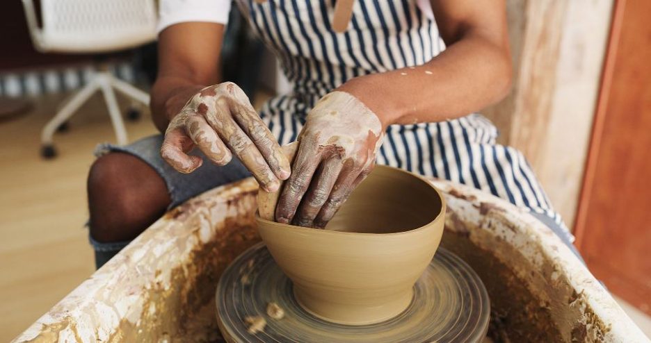 Close up of a woman in a striped apron creating a clay bowl on a wheel