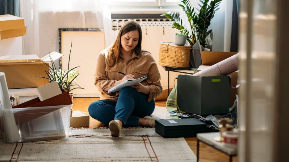Thoughful woman sitting on the floor of her home and writing notes while surrounded by boxes and belongings