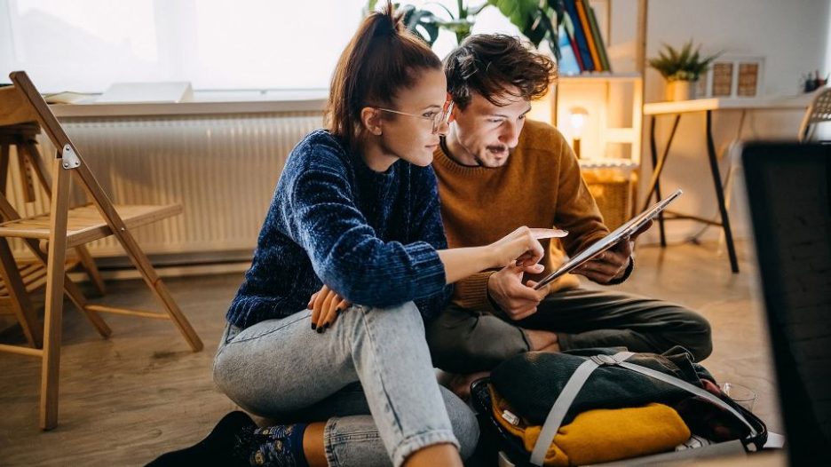 Couple seated on the floor of their home with a packaged suitcase, using a tablet together