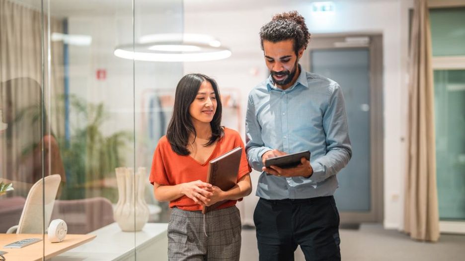 Two handsome, diverse colleagues are seen engaging in a productive conversation while utilizing wireless technology and a digital tablet in a modern office setting. Their smart casual attire adds to their professional appearance as successful employees.